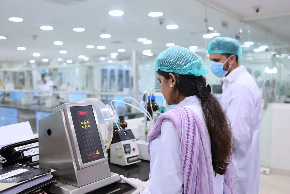 Techniciens en charlotte et blouse blanche travaillant dans un laboratoire de contrôle qualité pharmaceutique derrière des cloisons vitrées